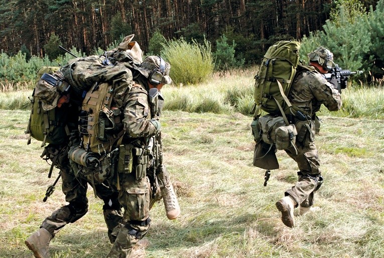 Oddział wychodzi z lasu. Żołnierze idą spokojnie z ciężkimi plecakami, z bronią gotową do strzału.