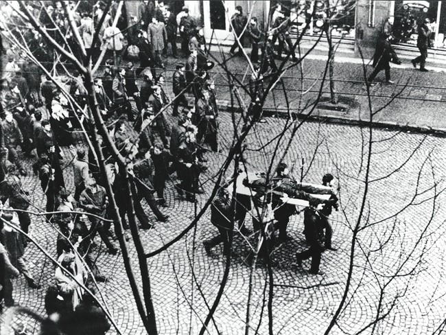 Gdynia, 17 grudnia 1970. Ulicą Świętojańską w kierunku centrum przechodzi pochód, demonstranci niosą na drzwiach zwłoki Zbigniewa Godlewskiego, który zginął w okolicy przystanku Gdynia -Stocznia, przeszyty serią z karabinu maszynowego