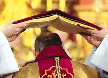  Otwarty ewangeliarz trzymany jest przez diakonów nad głową przyjmującego święcenia biskupie podczas całej modlitwy święceń