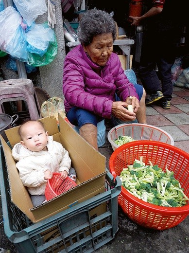 Ciepło ubrane dziecko w kartonie obok babci krojącej brokuły.
11.12.2017 Tajpej, Tajwan