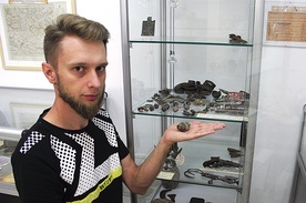 ◄	Sebastian Mielczarek obok półki z przedmiotami znalezionymi na terenie dawnego obozu jenieckiego. Na dłoni trzyma fragment zapalnika artyleryjskiego.