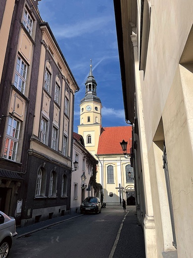 ▲	Świątynia z jednej strony jest otoczona wąskimi uliczkami wiodącymi na pszczyński rynek. Z drugiej strony rozciąga się kompleks słynnych ogrodów.